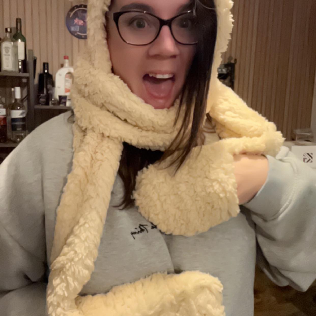 Picture of Chez A woman wearing glasses and a cozy, fluffy beige scarf and hoodie, smiling excitedly indoors with a background of bottles and wooden paneling.