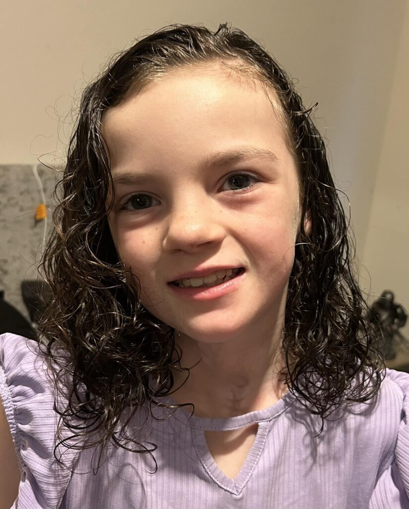 A young girl with wet, curly brown hair smiling at the camera, wearing a light purple top with a small cutout near the neckline. The background shows a kitchen setting with some appliances and a beige wall.