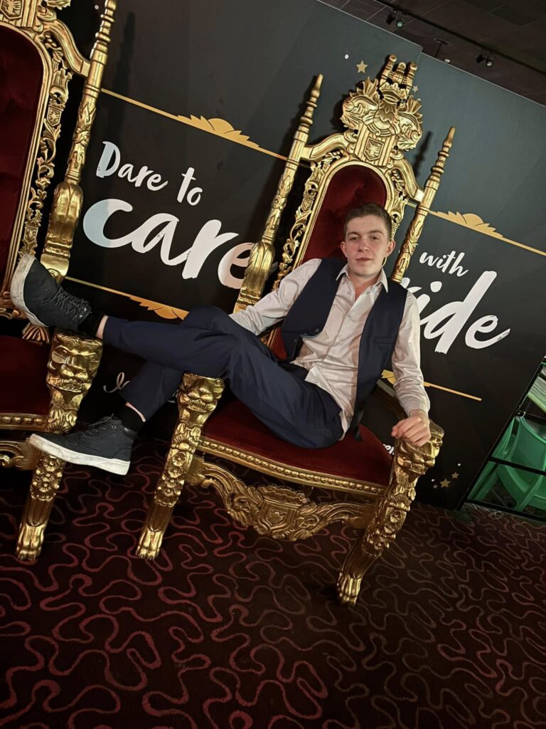 A young man sitting confidently on an ornate, gold-trimmed red throne with a backdrop that reads "Dare to care with pride." He is dressed in a white shirt, dark vest, and dark pants, with one leg crossed over the other and a relaxed expression.