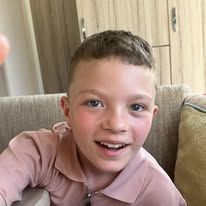 A young boy with short brown hair and blue eyes, smiling happily at the camera. He is wearing a pink collared shirt and sitting on a beige couch in a room with wooden cabinets in the background.