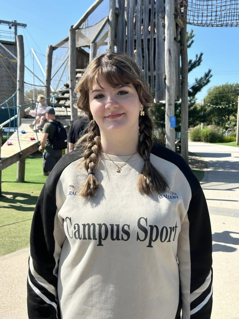 A young woman with braided brown hair, wearing earrings and a beige and black sweatshirt that says "Campus Sport," standing outdoors at a playground. She is smiling softly, with a wooden climbing structure and other children and adults in the background under a clear blue sky.