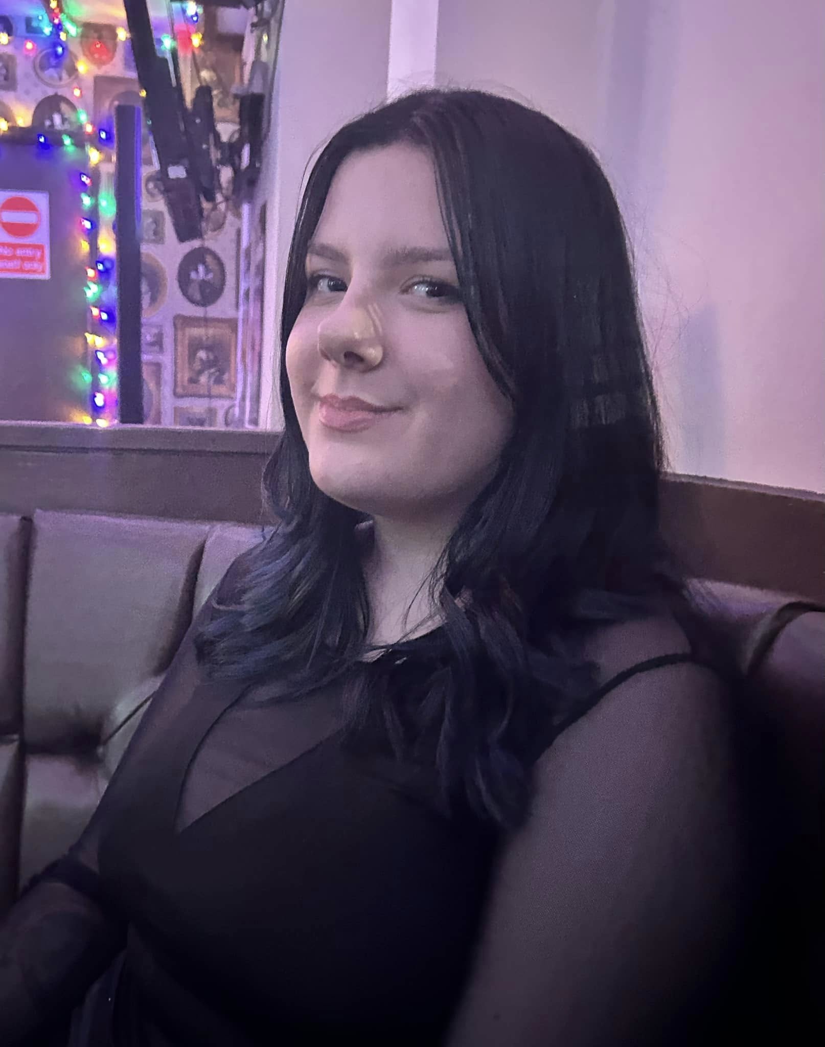 A woman with dark, wavy hair and fair skin, wearing a black sheer top, sitting on a cushioned bench in a dimly lit room with purple lighting. She is smiling softly and looking slightly to the side. In the background, colorful string lights and wall decorations are visible.